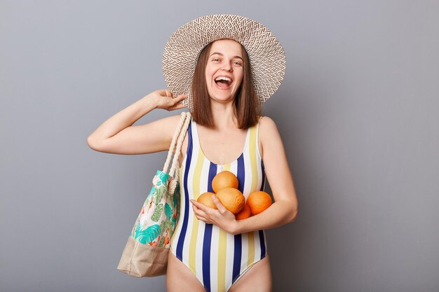 Summer traveling Smiling laughing young adult woman wearing striped swimsuit and straw hat holding fresh fruit enjoying resting having vacation isolated gray background