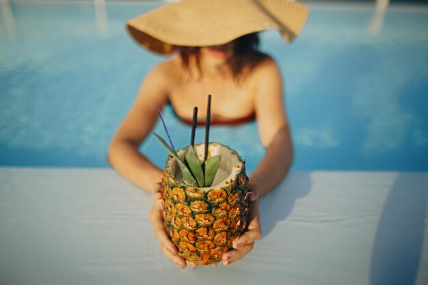 Summer vacation beautiful young woman in hat holding delicious cocktail in pineapple and relaxing in pool girl enjoying warm sunshine in pool on rooftop in luxury tropical resort top view