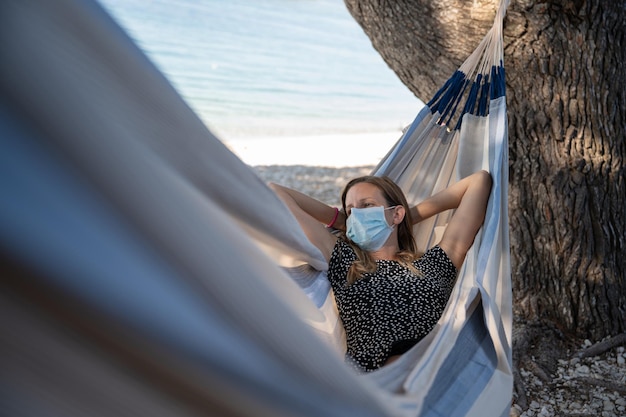 Summer vacation during coronavirus pandemic young woman lying in a hammock on the beach wearing a protective medical face mask