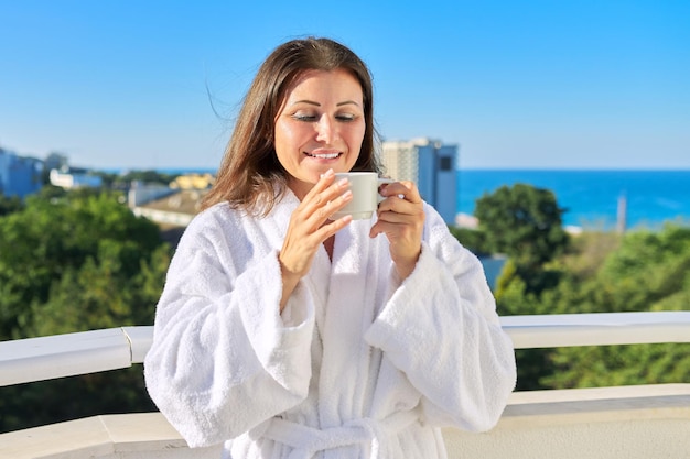 Summer vacation of happy mature woman. Middle-aged female in white bathrobe on balcony of hotel in seaside resort town. Enjoying cup of coffee, sun, beautiful seascape