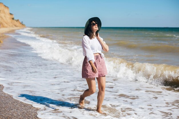 Summer vacation happy young boho woman walking in sea waves in sunny warm day at tropical island and blue sky space for text stylish hipster girl in hat relaxing on beach and smiling