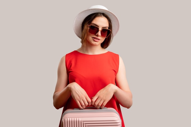 Фото Летние каникулы путешествие в отпуск улыбающаяся женщина в красном платье, солнцезащитные очки в белой шляпе с розовым чемоданом, смотрящая в камеру, изолированную на нейтральном пространстве для копирования туристическое агентство