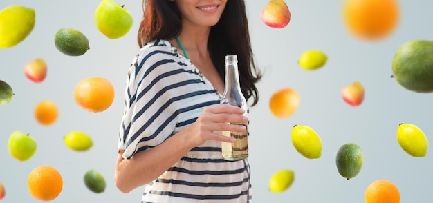 summer vacation, holidays, drinks and people concept - close up of smiling young woman drinking from bottle over fruits and berries on gray background