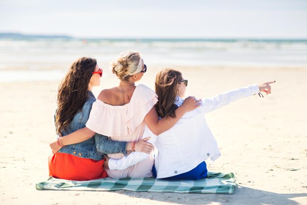 여름 휴가, 휴일, 여행 및 사람들의 개념 - 해변 담요에 앉아 손가락으로 무언가를 가리키는 젊은 여성 그룹