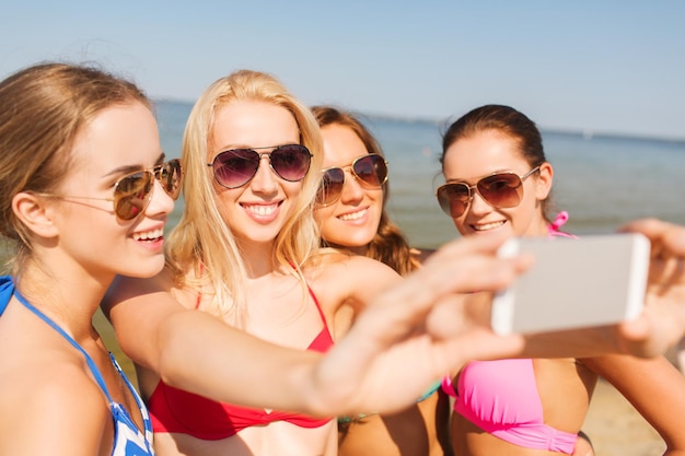 summer vacation, holidays, travel, technology and people concept- group of smiling young women on beach making selfie with smartphone over blue sky background