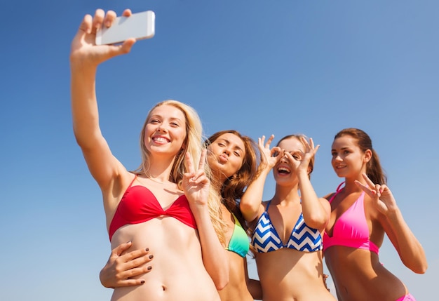 夏休み、休日、旅行、テクノロジー、人々のコンセプト-青い空を背景にスマートフォンで自分撮りを作るビーチで笑顔の若い女性のグループ