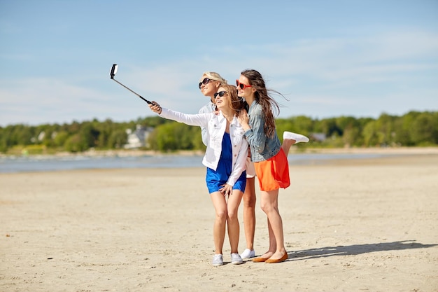 summer vacation, holidays, travel, technology and people concept- group of smiling young women taking picture with smartphone on selfie stick on beach