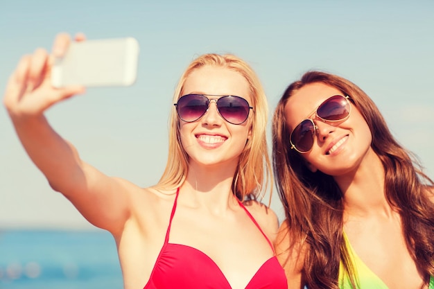 여름 방학, 휴가, 여행, 기술 및 사람 개념 - 해변에서 웃고 있는 두 젊은 여성이 푸른 하늘 배경에서 스마트폰으로 셀카를 찍고 있습니다.