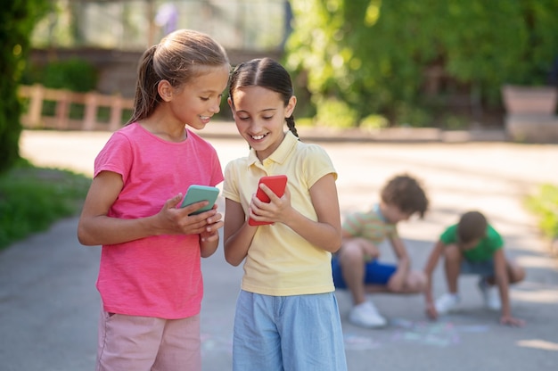 Summer vacation. Long-haired smiling good-looking two girls standing with smartphones and boys crouching drawing on asphalt behind