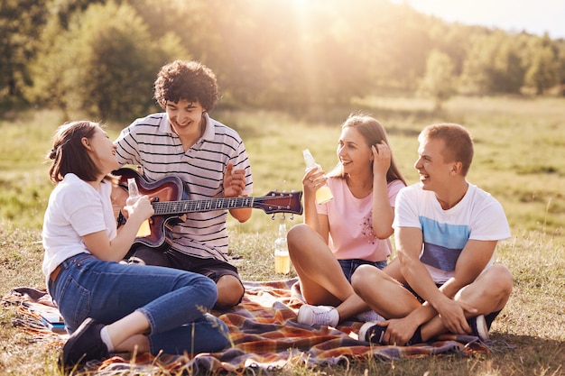 Summer, vacation, music and recreation time concept. Cheerful four friends or classmates have picnic outdoor, sing songs to guitar, drink energetic beverages, enjoys sunny warm day, pose on field