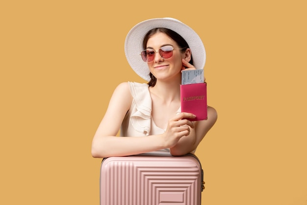 Summer vacation Passport control Holiday trip Flight checkin Happy woman sunglasses hat leaning suitcase holding foreign passport tickets looking at camera isolated on beige