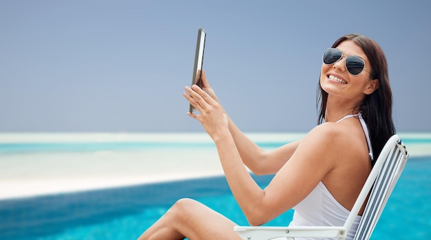 夏休み、観光、旅行、休日、人のコンセプト – ラウンジで日光浴をしているタブレットPCコンピュータを持つ笑顔の若い女性、ビーチとスイミングプールの背景に折りたたみ椅子