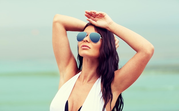 Concetto di vacanze estive, turismo, viaggi, vacanze e persone - giovane donna in costume da bagno con occhiali da sole sulla spiaggia