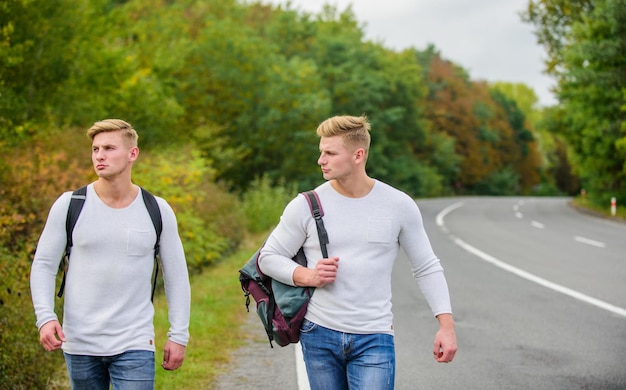 Vacanze estive viaggiatore turistico fermata automatica viaggiatori economici zaini da uomo che camminano insieme per strada gemelli che camminano lungo la strada avventura e scoperta uomo con zaino che fa l'autostop su strada