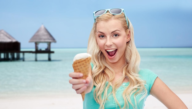 summer vacation, travel, tourism, junk food and people concept - young woman or teenage girl in sunglasses eating ice cream over beach on touristic resort background