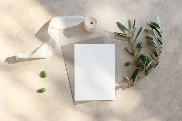 Summer wedding stationery mockup scene Blank greeting card invitation Craft envelope olive fruit branch and silk ribbon Elegant marble background in sunlight shadows Flat lay top view