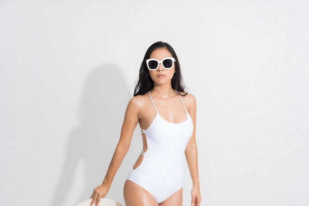 여름 여자 스튜디오 초상화입니다. 흰색 수영복 드레스를 입은 아시아 여성이 고립된 흰색 배경에 여름 패션으로 흰색 흰색 모자와 선글라스를 들고 서 있는 자세로 서 있습니다.