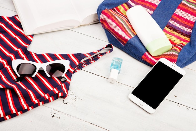 Фото Летняя женская мода с красочной полосатой сумкой, купальником, солнцезащитными очками для смартфона и книгой на белом деревянном столе