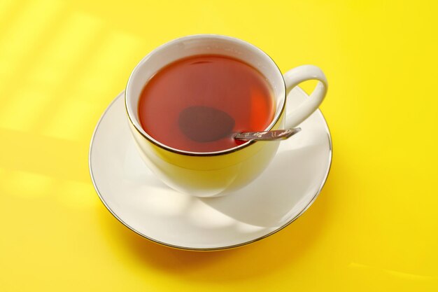 Photo sun shines over cup of hot tea with silver spoon on yellow board..