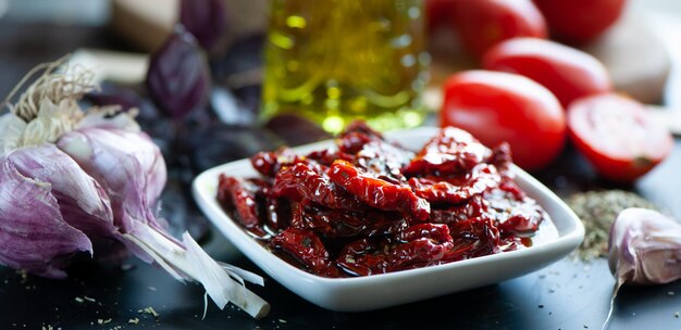 Sundried tomatoes in a bowl surrounded by ingredients spices garlic and basil to prepare Rustic style banner