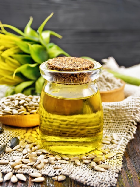 Sunflower oil in glass jar spoon with peeled seeds and flour in bowl on a burlap napkin a flower on dark wooden board background