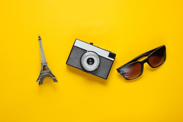 Sunglasses, retro camera, figurine of the Eiffel tower on yellow background. Travel background. Top view