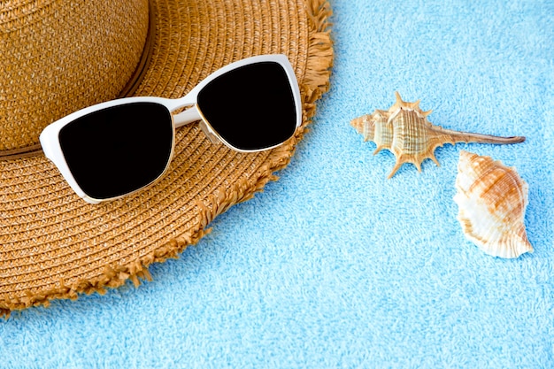 Sunglasses on a straw hat with seashells on a blue towel, summer concept.