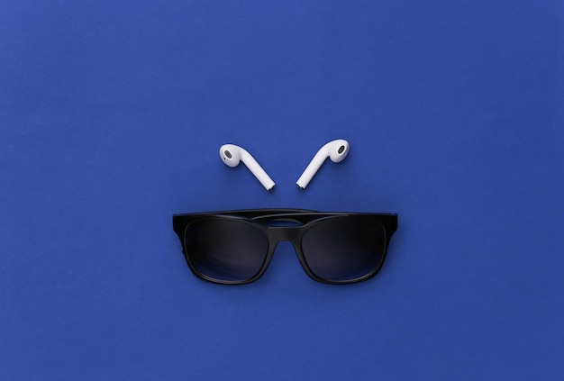 Sunglasses and wireless earphones on classic blue background. 