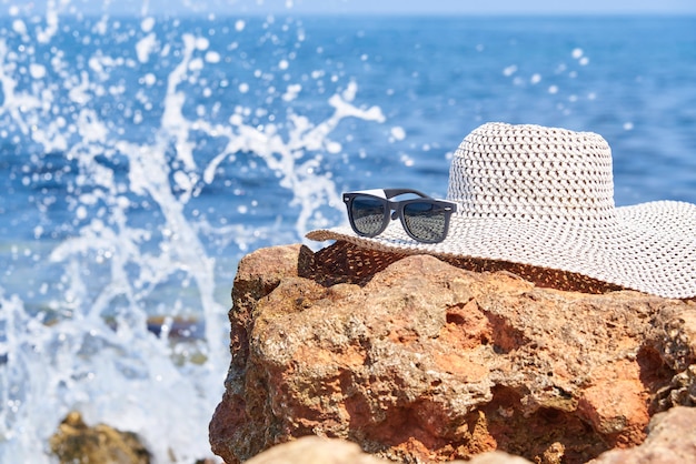 Sunglasses with a hat on a stone on a background of a sea with splashes of waves.