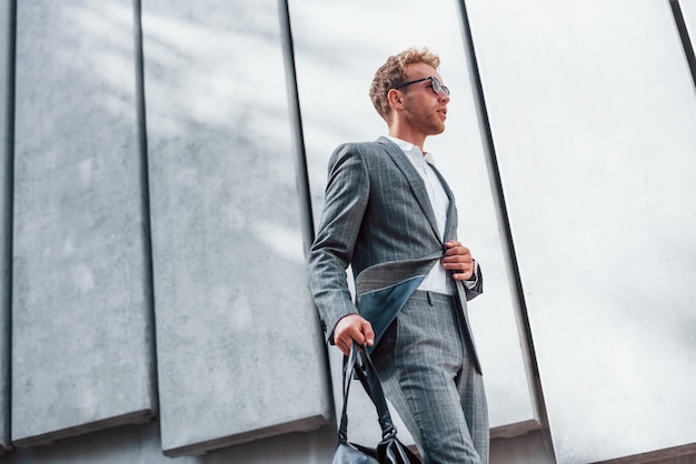 In sunglasses Young successful businessman in grey formal wear is outdoors in the city