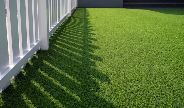 Sunlight and shadow of white wooden fence on green artificial\
turf surface in front yard of home