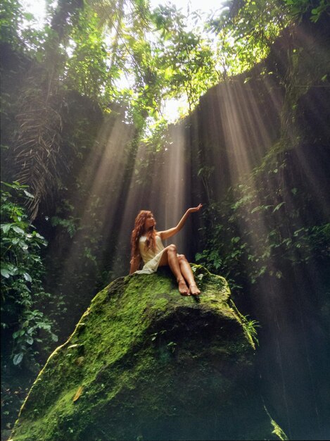 Солнечный свет проходит сквозь деревья, в то время как женщина сидит на моховой скале.