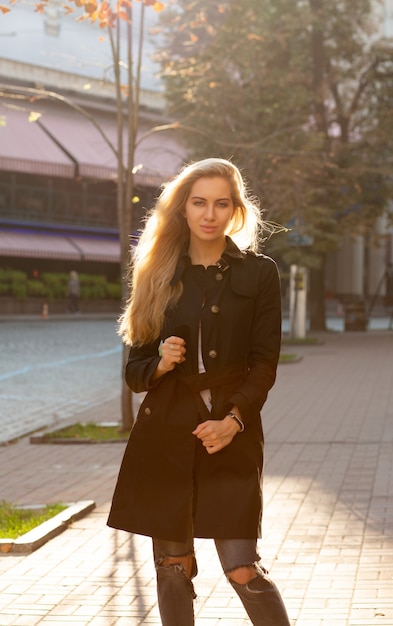 Sunny autumn portrait of wonderful blonde model wearing black coat and posing at the street