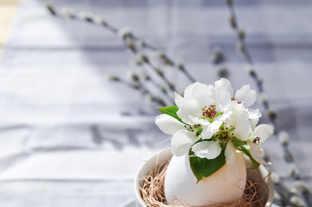 Sunny Easter-samenstelling van wilgentakken en natuurlijke bloemen in een wit ei in een cirkel op grijs tafelkleed.