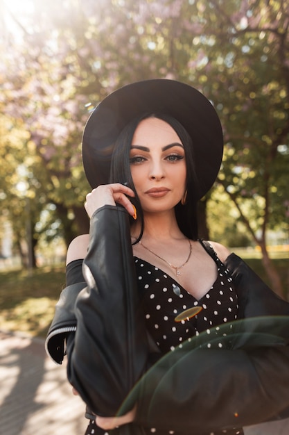 Sunny fresh portrait sexy woman in black beautiful dress in fashionable hat in vintage leather jacket in park on sunny bright day. Attractive sexy brunette girl enjoys sunlight on nature in spring day