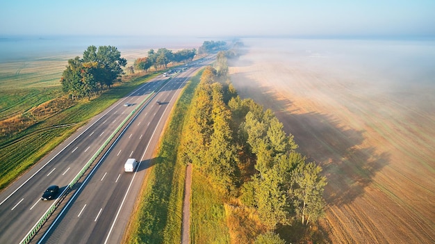 Солнечное волшебное осеннее туманное утро. Вид с воздуха на шоссе с автомобилями. Асфальтовая дорога покрыта туманом. Ранний туманный рассвет. Магистарал М1 в Беларуси