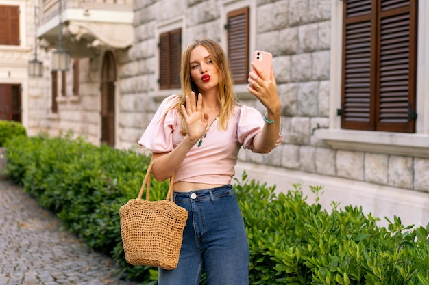 Photo sunny outdoor image of stylish blomde woman travel in europe spring summer vacation 70s outfit making selfie on the street holding smartphone