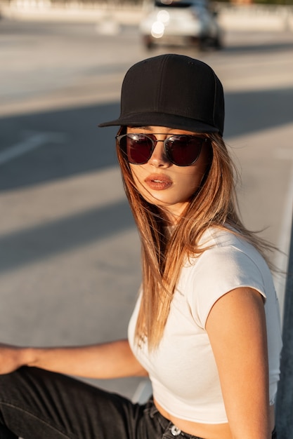 Sunny portrait cool beautiful young woman in sunglasses in fashionable black cap in white torn stylish t-shirt on parking lot at bright summer sunset. Fashion model swag girl rest on sun in city.