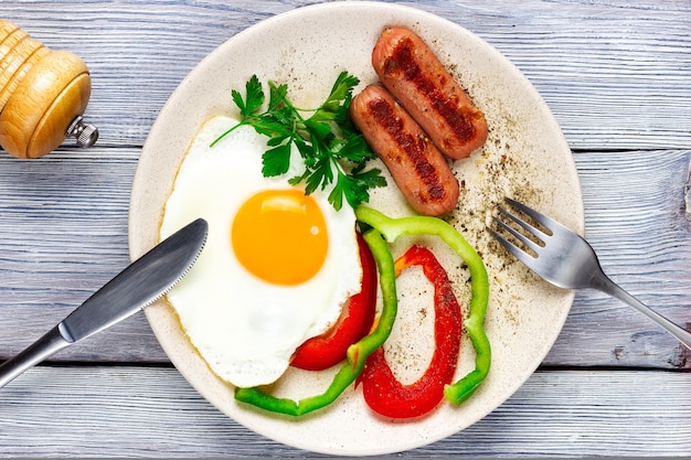 Sunny side up fried eggs with sausages and vegetables on a light wooden table fork and a knife top view