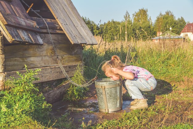 The Sunny summer day a child in a well draws water in a bucket and drinks from a metal mug