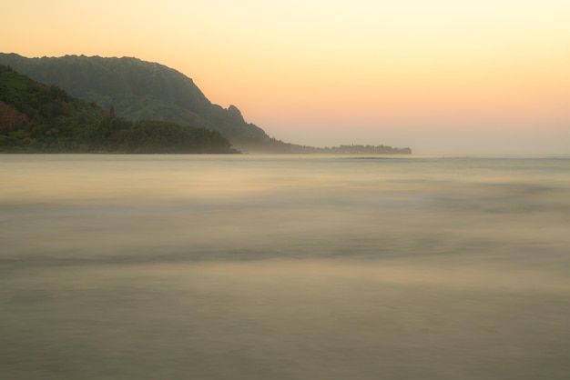 日の出はハナレイ湾の上の夜明けの空を照らし、ハナレイカウアイハワイの近くの背景にナパリ海岸があります