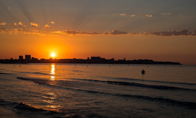 The sunrise seen from the postiguet beach in Alicante, with buildings and people against the light.