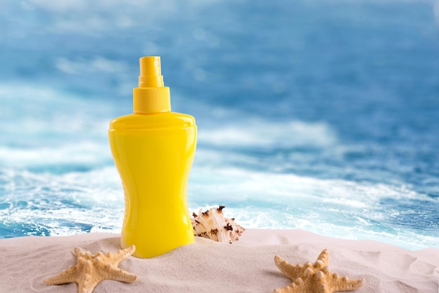 Sunscreen spray bottle on a sandy beach decorated with different seashells against sea waves background