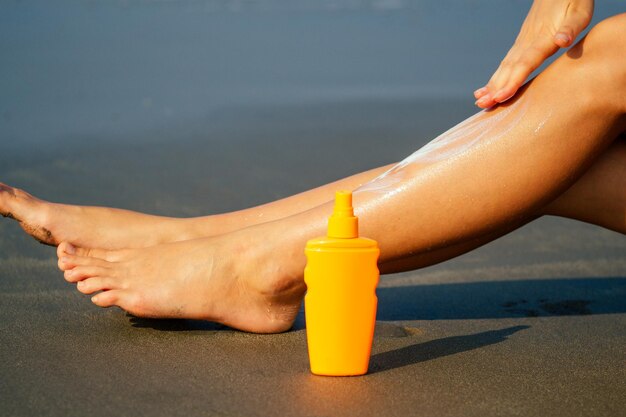 Sunscreen suntan lotion in spray bottle. young woman in\
spraying tanning oil on her leg from bottle. lady is massaging\
sunscreen lotion while sunbathing at beach summer vacation.