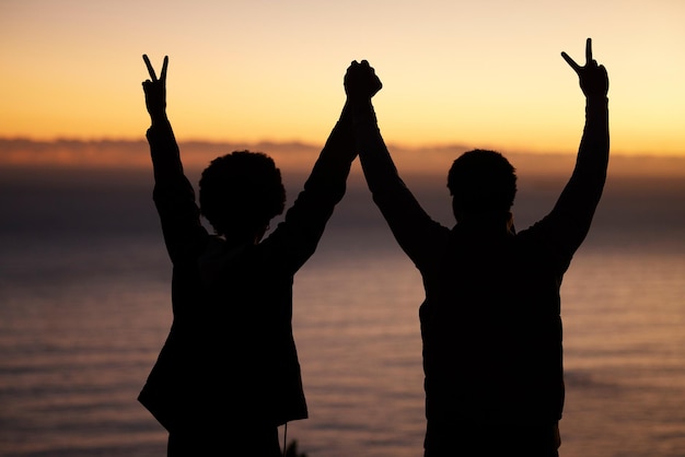 Sunset beach and silhouette of couple celebration success or winning and holding hands in shadow night or dark Sky ocean and winner people in rear with peace sign for sea hiking or travel goals