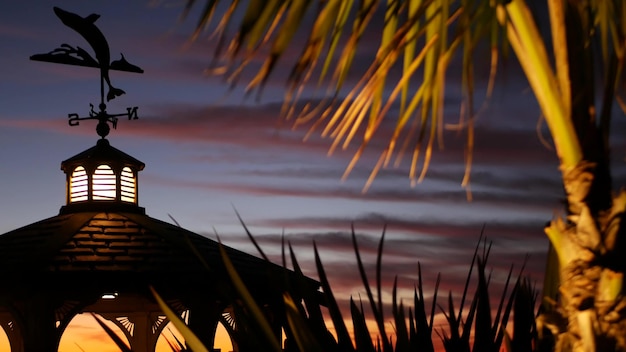 Sunset cloudscape and gazebo silhouette on beach, twilight\
dusk, clouds in dramatic purple pink sky, california coast. wooden\
romantic cosy alcove and palm tree. weather wind vane or roof\
weathercock.