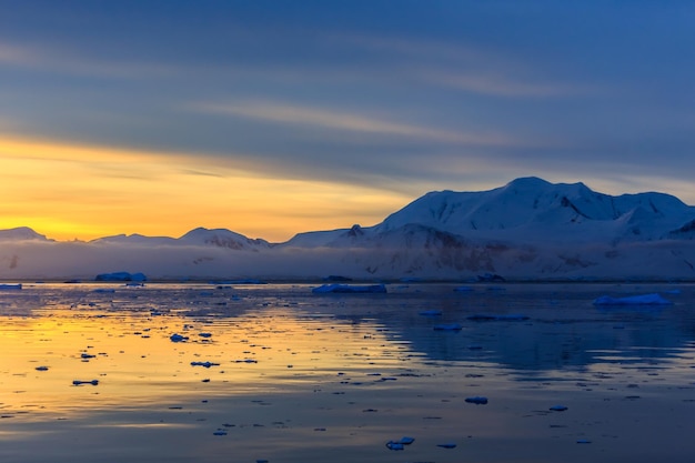 Lemaire Strait Antarctica의 백그라운드에서 산과 빙산이 있는 목가적인 석호 위로 일몰