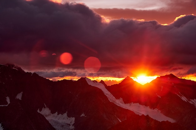 山の夕日。山の雪のピークと雲に赤い太陽の反射。アルタイ、ベルーハ地区