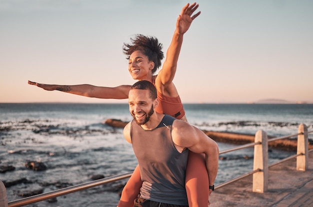 Sunset relax and couple piggyback by ocean enjoying holiday vacation and quality time on weekend Love freedom and happy black man and woman after exercise fitness workout and training by sea