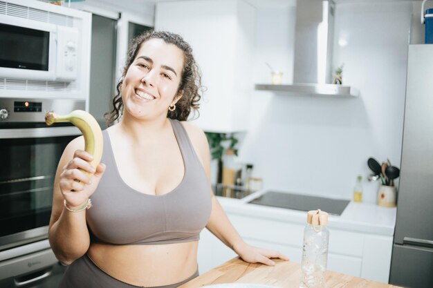 Super happy plus size curly woman smiling to camera while\
eating fruit at the kitchen home to lose weight training clothes\
getting fit for the summer conceptbeach body preparation healthy\
life